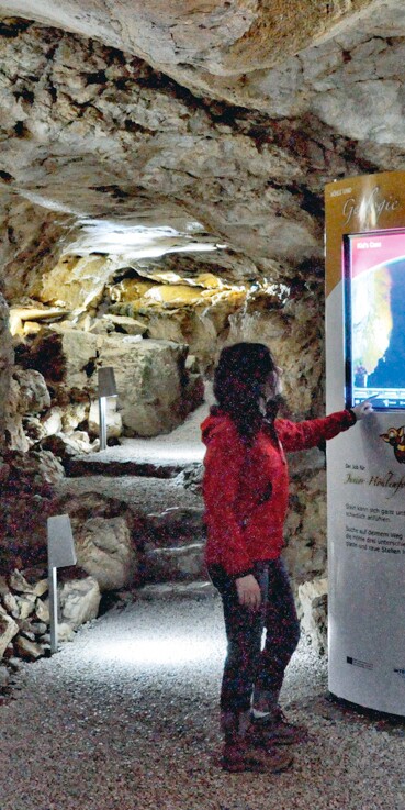 Eine Frau steht an einer interaktiven Informationstafel in der Wendelsteinhöhle. Die Höhlenwände werden mit Strahlern beleuchtet.