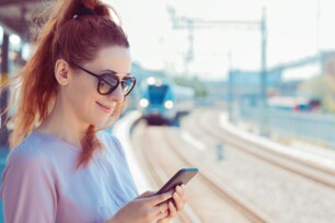 Eine Frau steht am Gleis und schaut lächelnd auf ihr Handy.