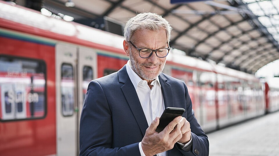 Ein Mann steht am Bahngleis und guckt auf sein Handy. Im Hintergrund steht eine bahn am Gleis.