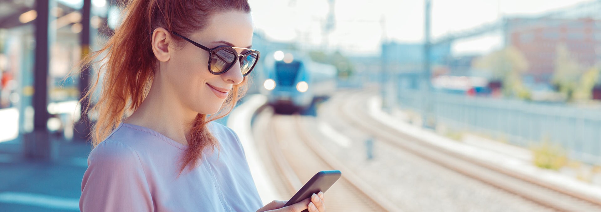 Frau mit Sonnenbrille am Bahngleis guckt auf das Handy