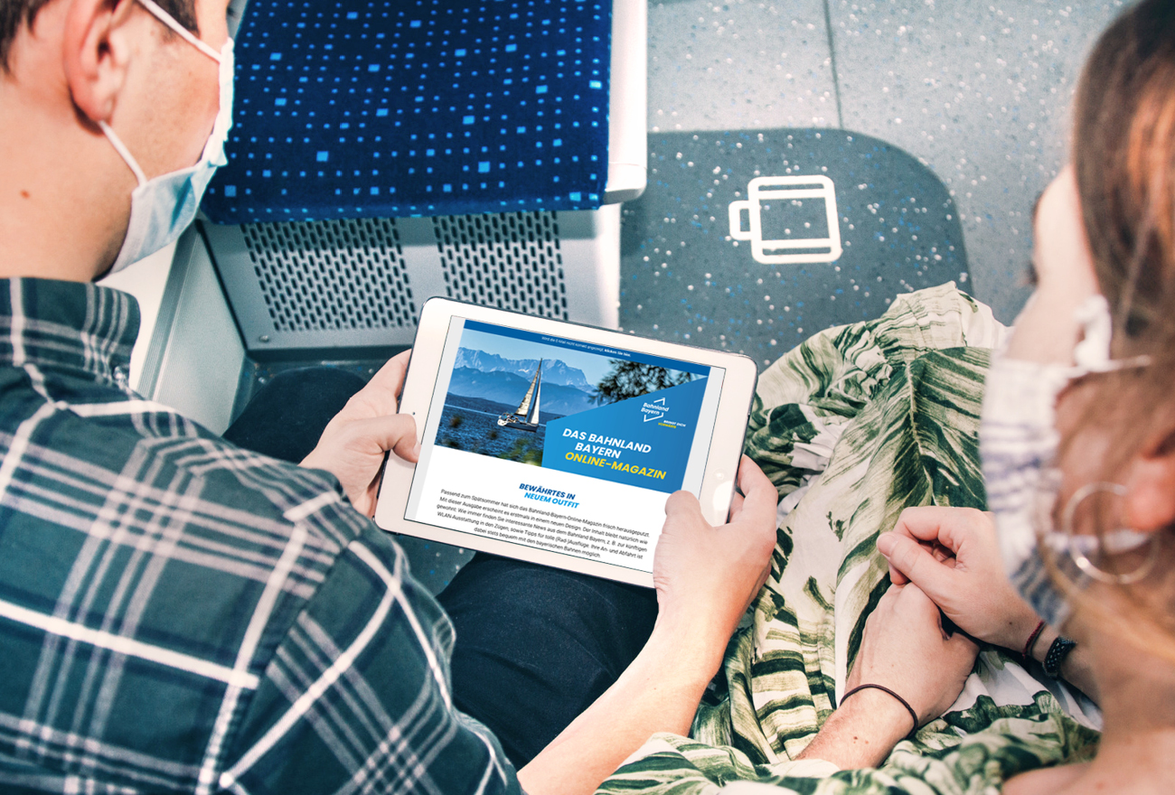 Ein Paar sitzt im Zug gemeinsam mit einem iPad. Es ließt das Bahnland-Bayern-Onlinemagazin.
